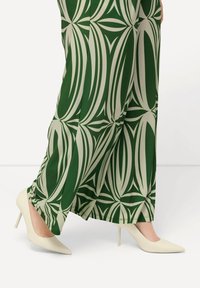 Breite Hosen mit grün-weißem Muster, die mit weißen spitz zulaufenden High Heels kombiniert sind. Der Stoff hat eine glatte Textur und ein geometrisches Design.