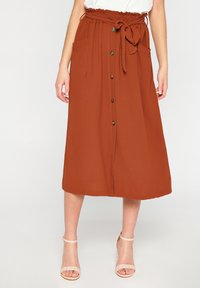 Gonna midi color ruggine con vita annodata, chiusura frontale con bottoni e tasche laterali. Tessuto leggero con una texture liscia.