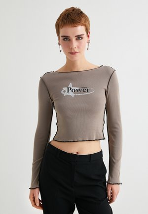 Long-sleeve cropped top in taupe with black stitching. Front features graphic text "Mystic Power" and star design. Smooth, stretchy fabric.