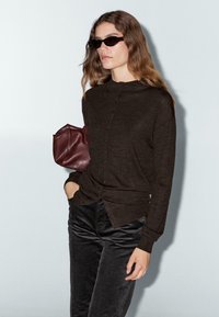Cardigan marrone a maniche lunghe con collo alto e chiusura con bottoni; abbinato a pantaloni in corduroy neri e una borsa bordeaux.