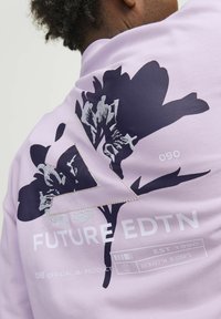 Sudadera lila con un gran estampado de flores oscuras, un acento triangular y texto en blanco que dice "FUTURE EDTN" con coordenadas geográficas abajo.