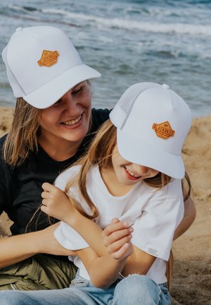 Hvide trucker caps med en brun læderlap, båret af en kvinde og en pige; sandstrand og vand i baggrunden. Afslappet tøj, lette teksturer.