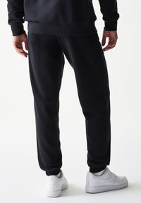 Μαύροι φόρμες με λεία υφή, ελαστικά μανσέτες και πλαϊνές τσέπες, σε συνδυασμό με λευκά αθλητικά παπούτσια. Απλός σχεδιασμός χωρίς διακοσμητικά στοιχεία.