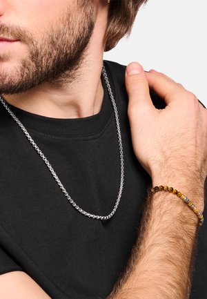 Silberne Kettenhalskette an einer Person, die ein schwarzes Hemd trägt; verfügt über ein glattes, rundes Design. Handgelenk mit einem Perlenarmband in Erdtönen geschmückt.