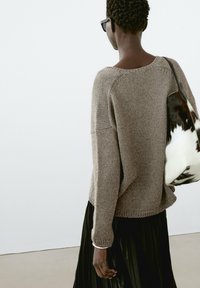 Pull marron en maille à coupe ample, avec des poignets côtelés et un ourlet arrondi, associé à une jupe plissée noire et un sac présentant un motif blanc et noir.