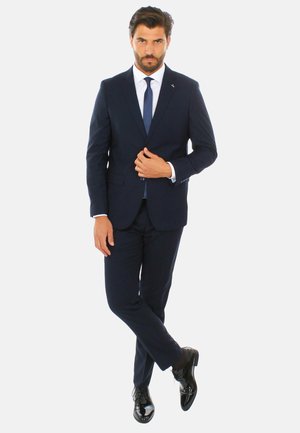Completo blu con un design su misura, caratterizzato da un risvolto a lancia, abbinato a una camicia bianca e una cravatta scura. Scarpe eleganti nere lucide completano l'outfit.