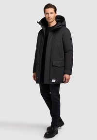 Muž v černém zimním kabátu s kapucí, černých kalhotách a černých botách, stojící na jednolitém světlém šedém pozadí.