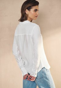 Hvid skjorte med lange ærmer, en afslappet pasform og en buet kant, parret med lyseblå jeans. Subtil tekstur og guldknapdetaljer.