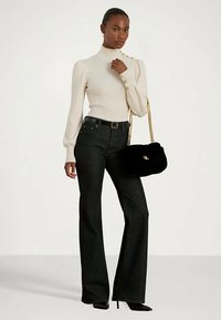 Žena u krem dugom rukavu s zlatnim gumbima, crnim visokim trapericama s raširenim nogavicama, crnim cipelama s visokom petom, drži crnu torbicu s zlatnom lancem.