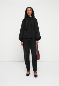 Γυναίκα που φοράει μαύρο πουκάμισο με μακριά μανίκια και φιόγκο, μαύρο τζιν, μαύρες γόβες και κρατάει μια μικρή μπορντό τσάντα.