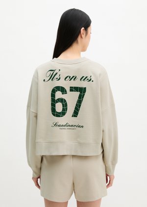 Kvinde med langt mørkt hår bærer beige sweatshirt og shorts, på bagsiden af sweatshirt står der "It’s on us. 67 Skandinavisk fodboldfællesskab."