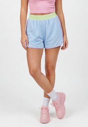 Kobieta ubrana w pastelowo niebieskie krótkie spodenki z zielonym paskiem, różowy krótki top, białe skarpetki i różowe masywne sneakersy, stojąca ze skrzyżowanymi nogami.