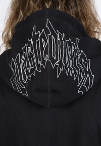 Schwarzer Hoodie mit einem weißen Grafikdesign auf der Rückseite. Das Material ist weich und hat eine glatte Textur. Kapuze ist angenäht, die Nähte sind sichtbar.