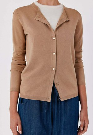 Persona che indossa un cardigan beige con bottoni sopra una camicia bianca e pantaloni di denim blu, mostrata dal collo alla vita su uno sfondo semplice.