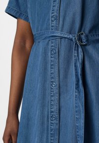 Dżinsowa sukienka z krótkimi rękawami, z przodu zapinana na guziki, z paskiem w talii z srebrnym pierścieniem oraz niebieską, lekko fakturowaną tkaniną.