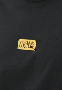 Svart t-skjorte med en gyllen metallisk etikett som leser "VERSACEJEANS COUTURE," med en glatt tekstur og rund hals.