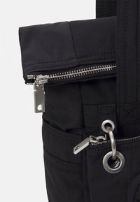 Section de sac en tissu noir avec deux fermetures éclair en métal argenté, dont une attachée à un anneau métallique circulaire par des sangles cousues.