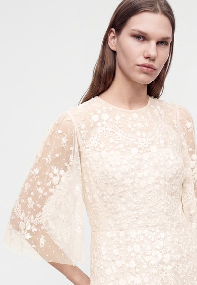 Jeune femme aux longs cheveux bruns portant une robe blanche transparente avec une broderie florale et des manches trois-quarts sur un fond uni.