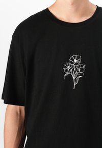 Sort bomulds T-shirt med en hvid floral linjetegning på brystet. Korte ærmer og en rund halsudskæring. Blød tekstur, afslappet design.