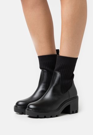 Bottes basses en cuir noir avec un dessus côtelé noir semblable à une chaussette et une semelle à talon épais. Texture lisse avec un bout arrondi et une semelle extérieure robuste.