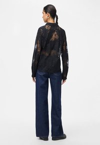 Woman with braided hair wears black sheer lace shirt, dark blue wide-leg jeans, black shoes, standing facing backwards on white background.