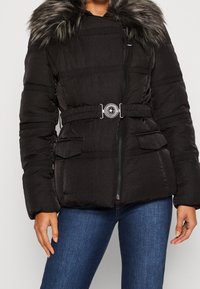 Chaqueta acolchada negra con capucha de piel sintética, cintura ceñida, cierre de cremallera y dos bolsillos frontales. Tejido texturizado con detalles de costuras horizontales.