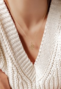 Collar de oro con un diseño de árbol, colgado de una delicada cadena, usado sobre un suéter de punto blanco texturizado.