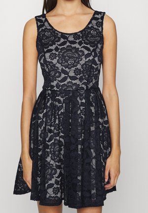 Robe noire sans manches en dentelle avec motif floral, taille cintrée et jupe évasée portée par une personne debout devant un fond uni.