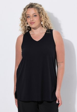 Donna con capelli biondi ricci indossa un top nero senza maniche e pantaloni neri, in piedi contro uno sfondo chiaro e uniforme con le mani rilassate.