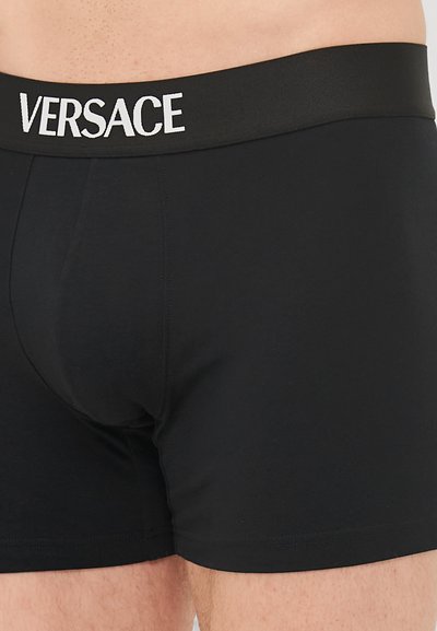 Črne bombažne boksarice z mehkimi elastiko, na kateri je v beli barvi napis "VERSACE", prilegajoč kroj in gladka tekstura.