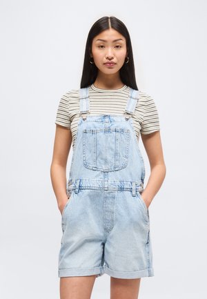 Νέα γυναίκα με ίσια μαύρα μαλλιά που φορά χρυσούς κρίκους σκουλαρίκια, ριγέ κοντομάνικο πουκάμισο και ανοιχτόχρωμα τζιν σόρτς με τιράντες, με τα χέρια στις τσέπες.