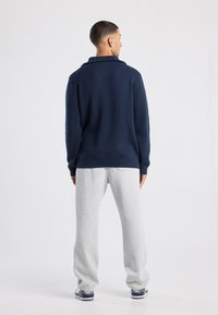 Man facing away wearing navy ribbed sweater, light gray sweatpants, and navy and white sneakers against plain white background.