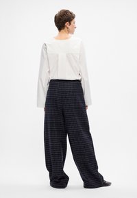 Bijela bluza dugih rukava s nabiranjem na leđima, u paru sa slobodnim tamnoplavićim kariranim hlačama. Glatkog materijala s opuštenim krojem.