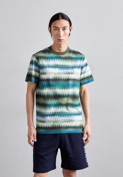Korte mouw katoenen t-shirt met een zigzagpatroon in groen, blauw en wit. Ronde halslijn, relaxte pasvorm, gecombineerd met donkere shorts.