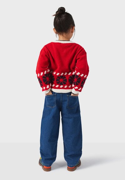 Roter Strickpullover mit weißen und schwarzen geometrischen Mustern, kombiniert mit blauen weit geschnittenen Jeans und braunen Schuhen. Gemütliche Textur und lebendiges Design.
