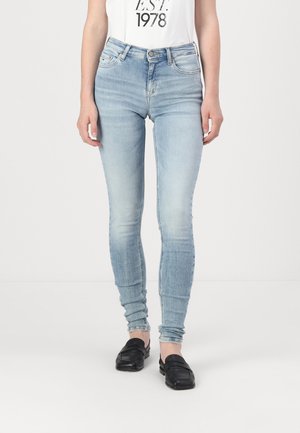 Mujer vestida con jeans ajustados de color azul claro, blusa blanca con el texto "EST! 1978" y mocasines negros, de pie frente a un fondo blanco liso.