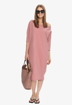 Kobieta w luźnej różowej sukience z odkrytymi ramionami, czarnych klapkach, dużych okularach przeciwsłonecznych, niosąca dużą brązową pikowaną torebkę.