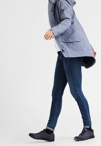 Chaqueta azul clara con capucha y bolsillos con cremallera sobre una camisa blanca, combinada con vaqueros azul oscuro y zapatillas negras con acentos morados.