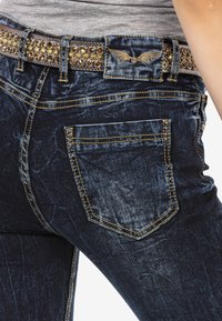 Dunkelblaue Jeans aus Denim mit einem detaillierten Bund, der mit goldenen Strasssteinen und dekorativen Nähten versehen ist. Enthält Gesäßtaschen mit Nieten.