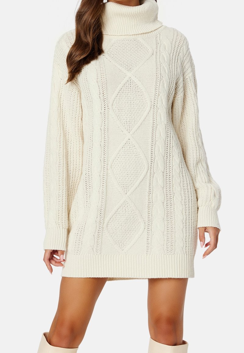 Femme portant une robe-pull col roulé ivoire à torsades, manches longues, avec un motif en losanges texturé à l'avant.