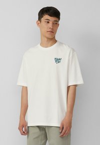 Jeune homme portant un t-shirt blanc ample avec le texte "Field Trip" sur la poitrine, associé à un pantalon vert clair, se tenant contre un fond gris.