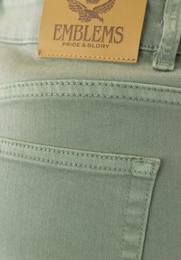 Tejido de denim verde con detalles de costura y un parche de cuero rectangular etiquetado "EMBLEMS" con "PRIDE & GLORY" grabado en marrón.