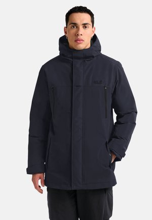 Mann trägt eine schwarze Winterjacke mit Kapuze, Reißverschlusstaschen auf der Brust und hat die Hände in den Seitentaschen, steht vor einem einfarbigen Hintergrund.
