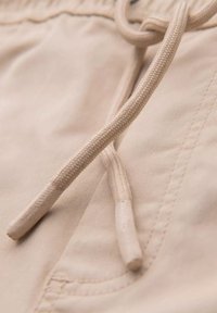 Primer plano de tejido beige con costura cosida y dos extremos de cordones, parte de una prenda con detalle de bolsillo.