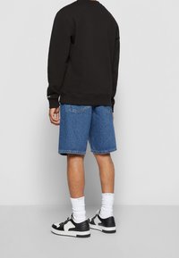 Personne portant un sweat-shirt noir, un short en denim bleu, des chaussettes blanches Calvin Klein et des baskets noires et blanches, debout devant un fond uni.