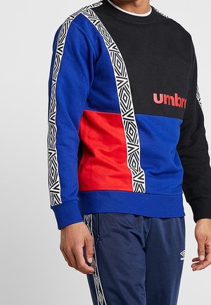 Mand iført en color-blocked sweatshirt i sort, blå og rød med mønstrede hvide striber og marineblå bukser med sidestriber.