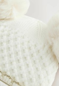 Weiße gestrickte Mütze mit einem strukturierten Muster, ausgestattet mit zwei hellen Fellpompons oben und einem silberfarbenen Akzentband am unteren Rand.