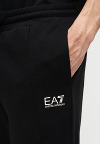 Το χέρι ενός άνδρα που ξεκουράζεται στην τσέπη μαύρου παντελονιού φόρμας Emporio Armani EA7 με εμφανές λευκό λογότυπο στον μηρό.