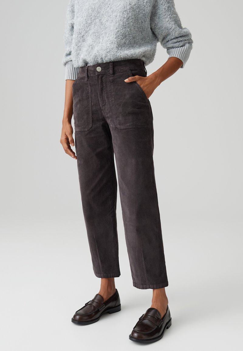 Personne portant un pantalon en velours côtelé marron foncé avec de grandes poches devant, un pull gris pelucheux et des mocassins marron foncé, debout sur un fond uni.