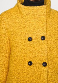 Cappotto di lana gialla a trama con colletto alto, dotato di due grandi bottoni neri e un sottile motivo a quadretti.